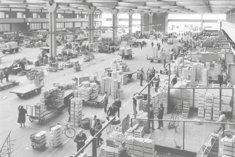 Fotografia d’epoca in bianco e nero del mercato ortofrutticolo al coperto con operatori e casse di prodotti